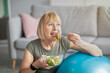 Leinwandbild Motiv Sports and nutrition concept. Smiling senior lady leaning on fitness ball, eating fresh vegetable salad at home