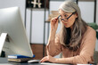 © Kateryna - Shocked excited influential mature gray-haired asian, business woman, manager, is sitting at computer, received an unexpected message, looks surprised at computer screen, takes off glasses