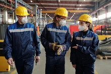 Group of workers or engineers in protective workwear and respirators moving along workshop with equipment and consulting