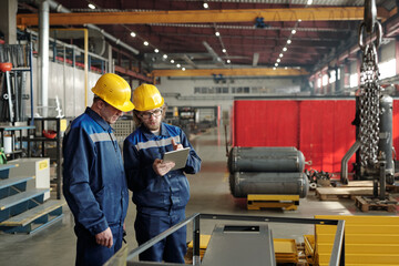 Canvas Print - Two male engineers in workwear standing in the middle of large workshop or plant and discussing characteristics of new industrial equipment