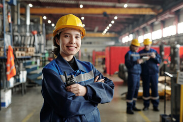 Poster - Young smiling female worker of modern industrial plant or factory in workwear and protective helmet standing in large workshop