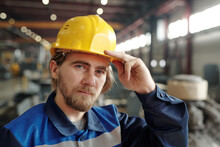 Young Serious Male Engineer Or Factory Worker In Yellow Hardhat Standing In Front Of Camera And Looking At You Against Workshop Environment