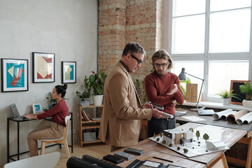 Sticker - Young male architect and his mature colleague in smart casualwear discussing parts of model of new house and yard at working meeting