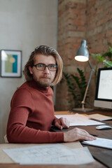 Wall Mural - Young bearded male architect in eyeglasses and casualwear looking at you while sitting by table in front of camera and working over new project