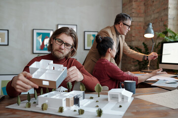 Poster - Young bearded male architect in eyeglasses holding part of new house model against his colleagues discussing sketch on computer screen