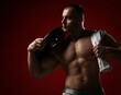 © Dmitry Lobanov - Portrait of muscular man, bodybuilder standing shirtless with naked chest holding towel and weight for barbell on shoulder and looks aside over red background