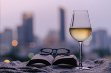 A Glass Of White Wine With Book And Spectacles Put On Bed With Colorful City Bokeh Lights Background. Stay Home Concept.