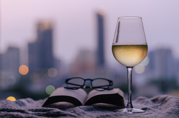 A glass of white wine with book and spectacles put on bed with colorful city bokeh lights background. Stay home concept.