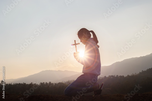 Silhouette of human kneeling down praying and holding christian cross for worshipping God at sunset background. Christian, Christianity, Religion copy space background.
