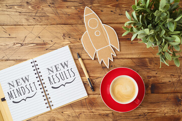 New mindset and new results text on notebook on wooden table with coffee cup and paper rocket. Business concept of positive thinking and motivation