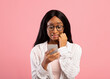 Oops. Embarassed black woman looking at mobile phone screen in confusion on pink studio background