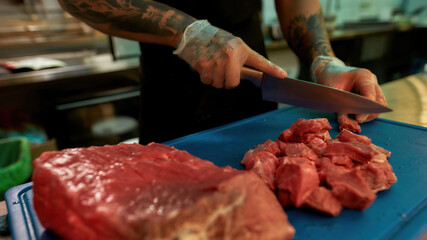 Wall Mural - Close up of hands of male cook wearing protective gloves chopping raw pork meat on a plastic board while working in commercial kitchen