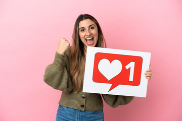 Wall Mural - Young caucasian woman isolated on pink background holding a placard with Like icon and celebrating a victory