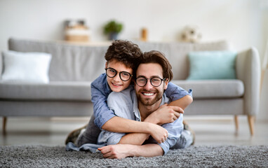 Happy father and son having fun at home. Dad lying on carpet carrying boy on back and smiling together to camera