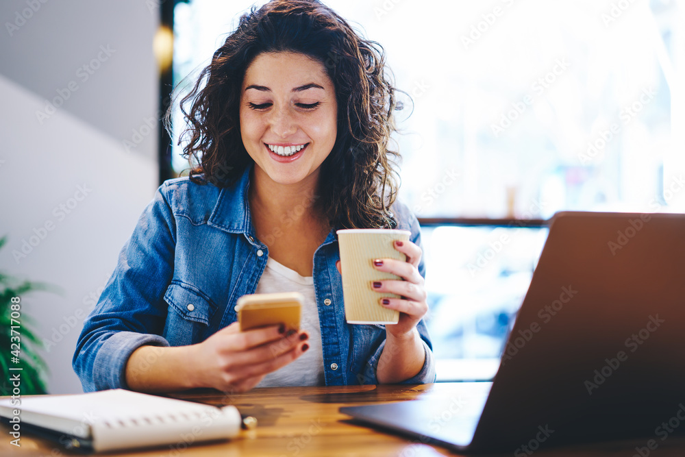Happy hipster girl with coffee to go using cellular device for online messaging and chatting in social media, cheerful female blogger with takeaway cup connecting to 4g for browsing website - obrazy, fototapety, plakaty 