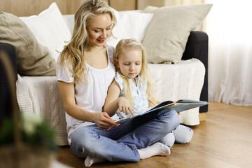 Happy family. Blonde young mother reading a book to her cute daughter while sitting at wooden floor. Motherhood concept