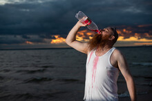A Humorous Portrait Of A Brutal Man Pouring Soda From A Bottle On The Beach At Sunset