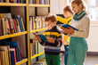 © Iryna - Cute different age pupils, two boys and girl looking for books in library at the elementary school. Three children standing near bookshelves and reading together at school's library.