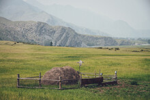Beautiful Foggy Mountain Landscape With Scarecrow On Wooden Fence Around Haystack On Background Of Mountains And Rocks Silhouettes In Fog. Stack Of Hay Is Surrounded By Wood Fence In Vintage Tones.