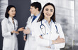© rogerphoto - Smiling intelligent woman-doctor is standing with arms crossed in a clinic, together with her colleagues at the background. Portrait of physicians at work. Perfect medical service in a hospital