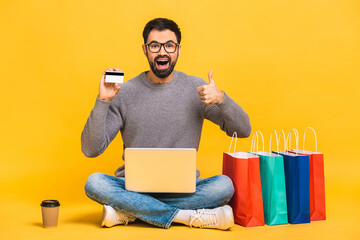 Shopping time! Portrait of happy young bearded man with shopping packages holding laptop computer while sitting on floor isolated on yellow background. Using credit card.