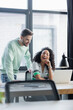 © LIGHTFIELD STUDIOS - young businessman standing near african american manager laughing near laptop, blurred foreground