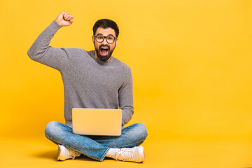 Successful winner! Portrait of a happy young bearded man using laptop and celebrating success or victory isolated over yellow background.