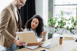 © LIGHTFIELD STUDIOS - Smiling businessman holding paper folder near african american colleague with gadgets in office