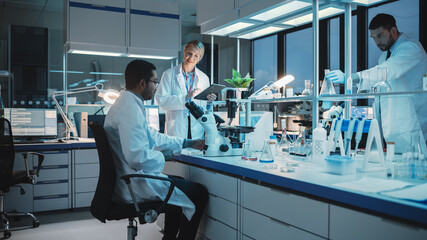 Wall Mural - Female and Male Medical Research Scientists Have a Conversation While Conducting Experiments Under a Microscope, Writing Analysis Results on a Tablet Computer. Modern Biological Science Laboratory.