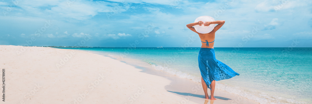 Beach vacation happy woman walking on summer travel Caribbean holiday with arms behind head wearing sun hat and sarong skirt. Ocean panoramic banner background. Elegant lady tourist. - obrazy, fototapety, plakaty 