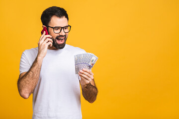 Wall Mural - Happy winner! Excited young bearded man in casual holding lots of money in dollar currency and cell phone in hands isolated over yellow wall. Using mobile phone.