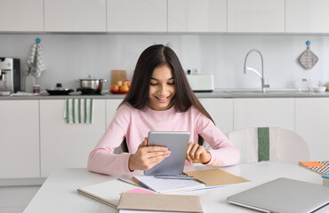 Indian kid school girl distance learning online at home using digital tablet. Happy child teen student remote studying holding pad computer homeschooling doing homework sitting at kitchen table.