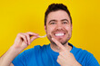 © Roquillo - young handsome caucasian man wearing blue t-shirt against yellow background holding an invisible aligner and pointing to her perfect straight teeth. Dental healthcare and confidence concept.