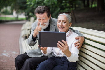 Beautiful aged couple in stylish clothes having video conversation on digital tablet while relaxing at green park. Concept of people and technology.