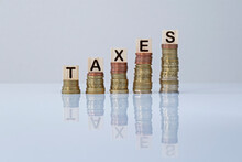 Word "TAXES" On Wooden Blocks On Top Of Ascending Stacks Of Coins Against Gray Background. Concept Photo Of Increase Of Taxes, Economy, Business And Finance.