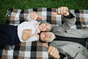 Stylish senior couple smiling and showing thumbs up while lying on soft checkered blanket. Happy aged man and woman spending time together on fresh air.