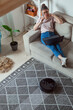 © perfectlab - Robot vacuum cleaner cleaning carpet, woman using laptop sitting