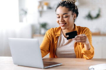 Happy young african american woman stylishly dressed, paying for online purchases, uses a laptop, holding a banking card in hand, typing credit card information on the keyboard and smiling