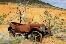 Old Vintage Rusted Car With Dead Tree Growing Out Of The Centre Of It. There Is Orange Hill With Dry Grassy Vegetation Behind.