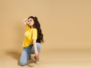 Young beautiful girl with long curly hair in a yellow t-shirt stands on one knee on a pastel orange studio background.