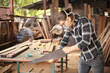 © Akarawut - Young Caucasian carpenter man is sawing plank of wood in his own garage style workshop for hobby with copy space