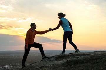 Wall Mural - Man and woman hikers helping each other to climb stone at sunset in mountains. Couple climbing on high rock in evening nature. Tourism, traveling and healthy lifestyle concept.