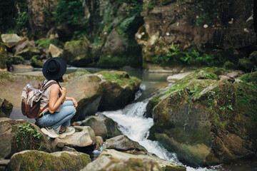 Wall Mural - Stylish woman traveler with backpack sitting at river in mountains, relaxing. Travel and wanderlust