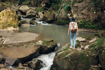 Wall Mural - Travel and wanderlust. Stylish woman traveler with backpack standing at river in mountains, hiking