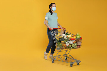 Poster - Woman with protective mask and shopping cart full of groceries on yellow background