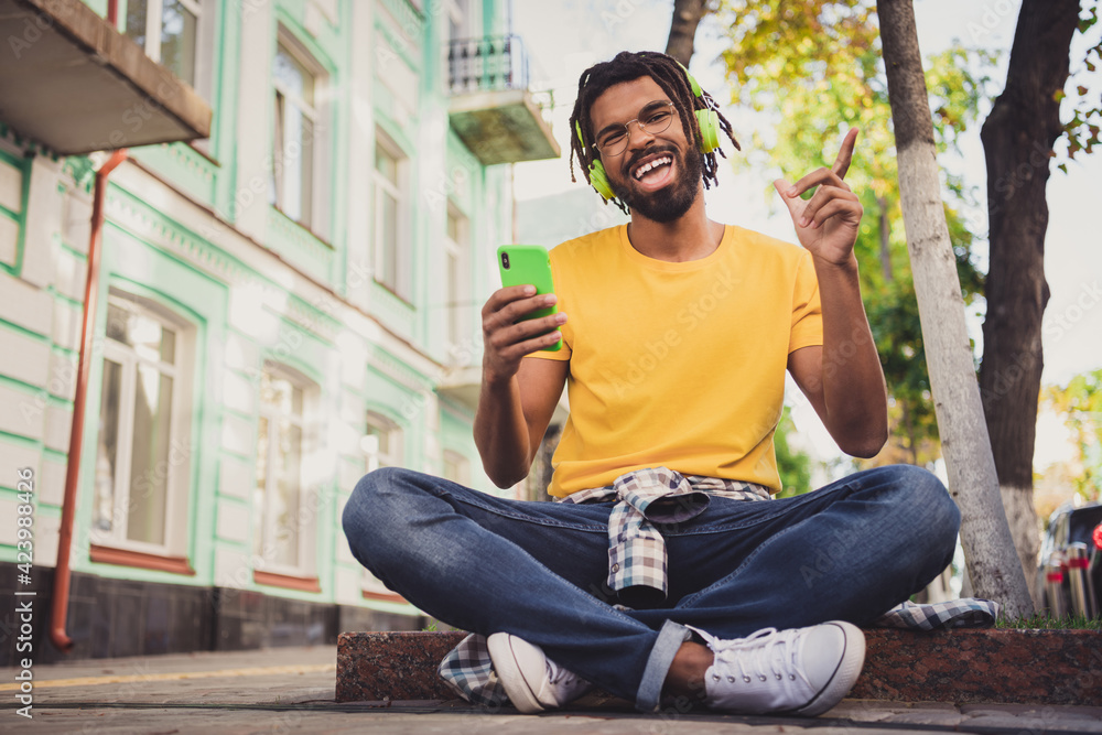 Photo portrait of guy sitting on the ground enjoying music with cellphone smiling on city street in summer - obrazy, fototapety, plakaty 