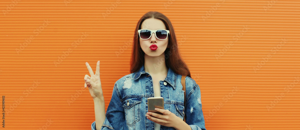 Portrait close up of young woman with smartphone wearing a denim jacket posing on an orange background - obrazy, fototapety, plakaty 