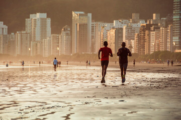 Two men running together on the beach at the end of a golden afternoon in Santos. In the background, people, buildings by the sea and on the hill.