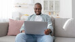 © Prostock-studio - Happy middle-aged black man using laptop while resting on sofa in living room, panorama
