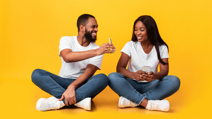 Black couple using mobile phones, sitting on floor at studio